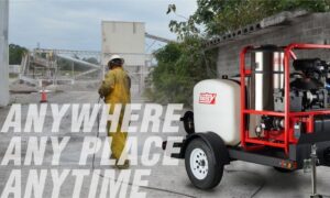 Worker using Hotsy pressure washer at industrial site with “Anywhere Anytime” slogan.