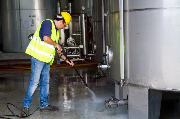 Worker using an industrial pressure washer to clean manufacturing equipment and facility floors inside an industrial plant.